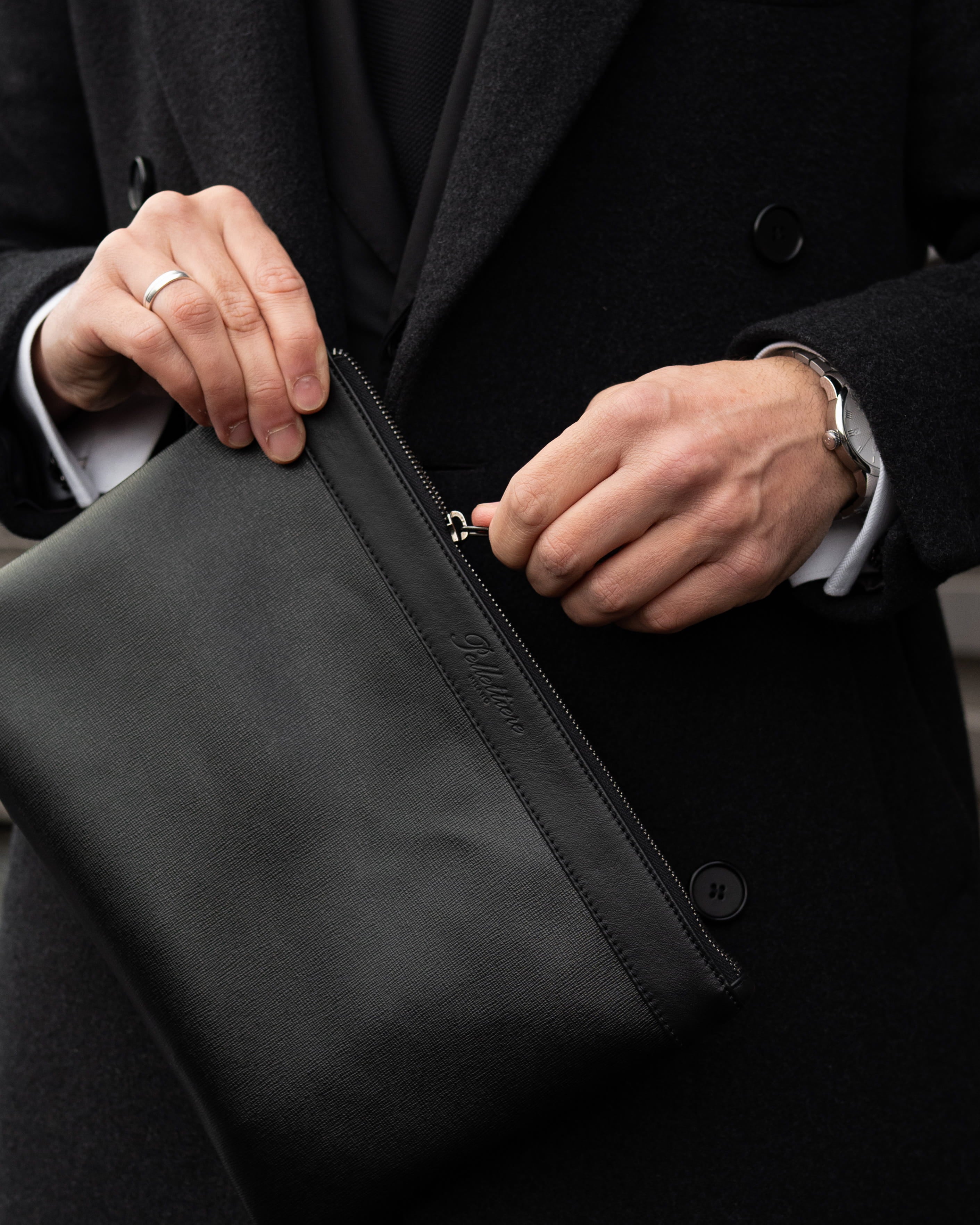 Nahaufnahme eines Mannes im dunklen Mantel, der eine schwarze Leder Tasche (Pouch) von Pellettiere Milano öffnet – elegantes Kartenetui Accessoire.